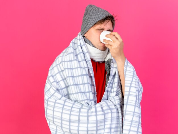 Jovem bonito loira doente com chapéu de inverno e lenço embrulhado em xadrez bebendo uma xícara de chá com os olhos fechados, isolado em um fundo carmesim com espaço de cópia