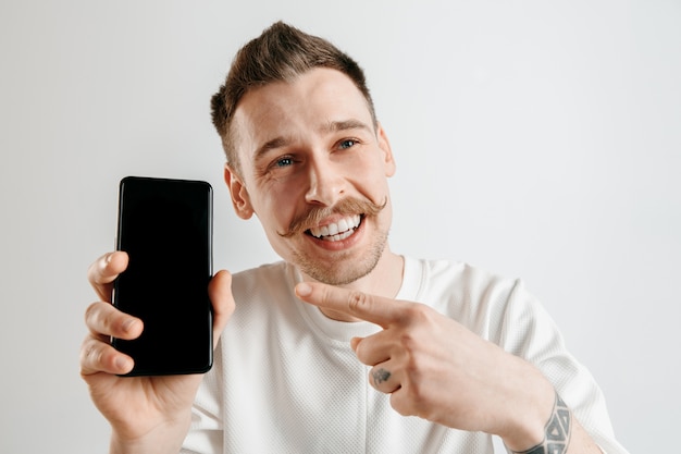 Jovem bonito mostrando a tela do smartphone em um espaço cinza com uma cara surpresa