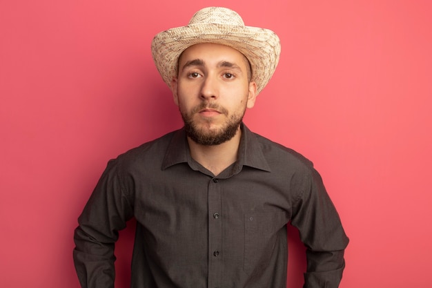 Jovem bonito olhando para a frente usando camiseta preta e chapéu isolado em rosa