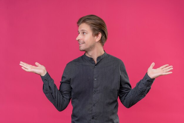 Jovem bonito parecendo confuso abrindo os braços sorrindo sem resposta em pé sobre a parede rosa