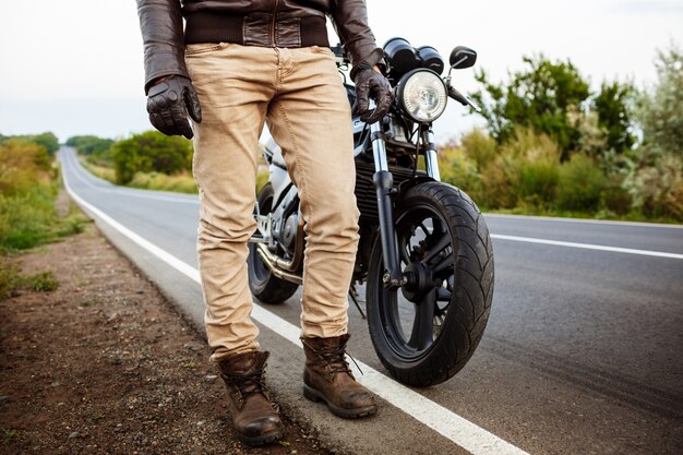 Jovem bonito posando perto de sua moto na estrada rural.