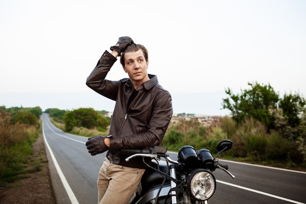 Foto grátis jovem bonito posando perto de sua moto na estrada rural.