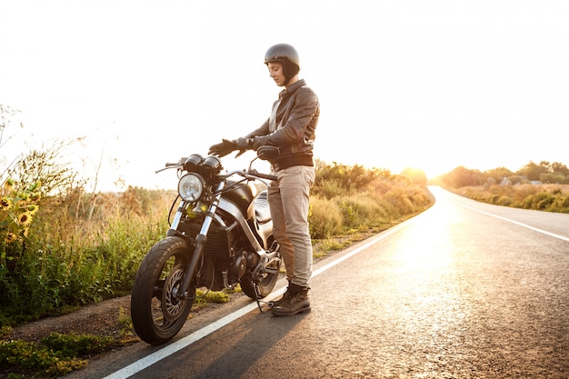 Jovem bonito posando perto de sua moto na estrada rural.