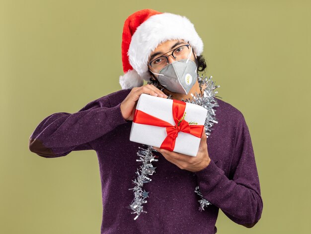 Jovem bonito satisfeito com chapéu de Natal e máscara médica com guirlanda no pescoço e segurando uma caixa de presente isolada em fundo verde oliva