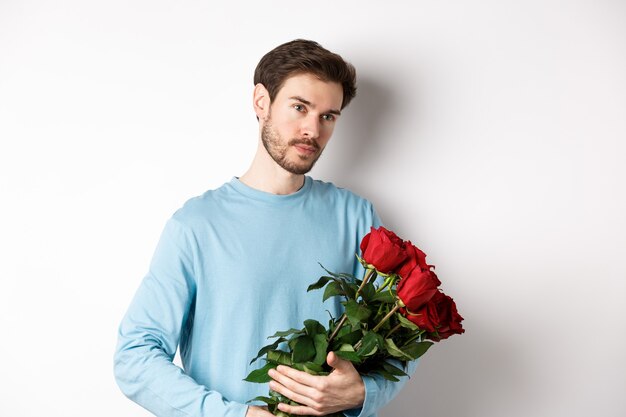 Jovem bonito segurando lindas rosas vermelhas para sua amante no dia dos namorados, parecendo pensativo, em pé sobre um fundo branco