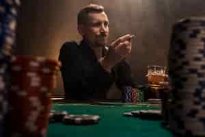 Foto grátis jovem bonito sentado atrás da mesa de pôquer com cartas e fichas. em primeiro plano, pilhas de fichas na mesa de pôquer em um quarto escuro cheio de fumaça de cigarro.