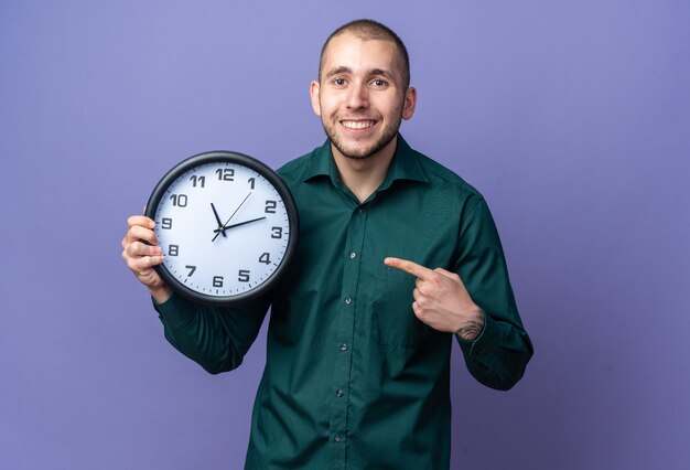 Jovem bonito sorridente com camisa verde segurando e apontando para o relógio de parede