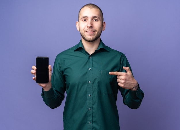 Jovem bonito sorridente de camisa verde segurando e apontando para o telefone