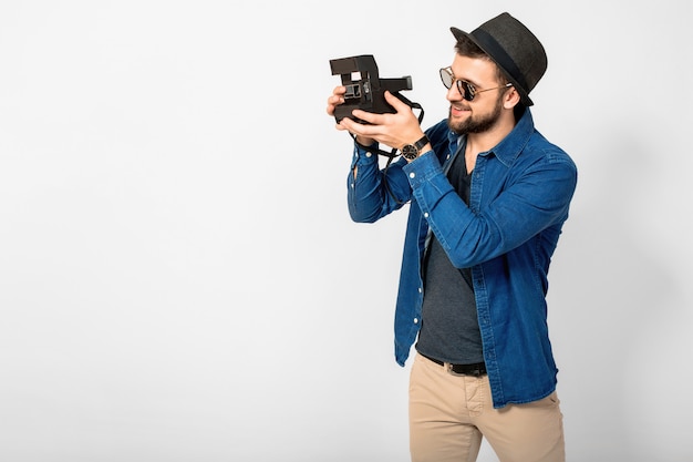 Jovem bonito sorridente homem feliz segurando uma câmera fotográfica vintage isolada no fundo branco do estúdio, vestindo camisa jeans, chapéu e óculos escuros, fotógrafo viajando e tirando fotos