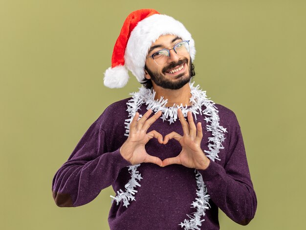 Jovem bonito sorridente usando chapéu de Natal com guirlanda no pescoço e mostrando gesto de coração isolado em fundo verde oliva