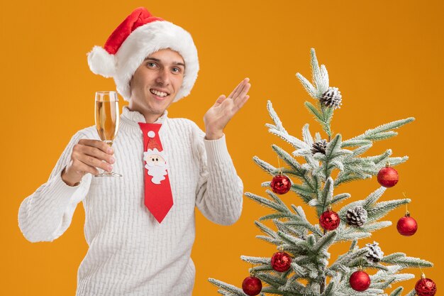 Jovem bonito sorridente usando chapéu de natal e gravata de papai noel em pé perto de uma árvore de natal decorada segurando uma taça de champanhe, olhando mostrando a mão vazia isolada na parede laranja