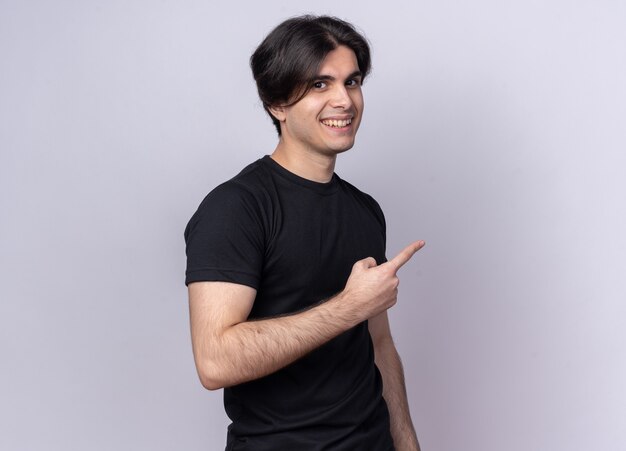 Jovem bonito sorridente usando uma camiseta preta apontando para o lado isolado na parede branca com espaço de cópia
