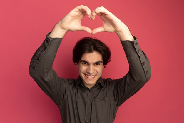 Jovem bonito sorridente usando uma camiseta preta e mostrando um gesto de coração isolado na parede rosa