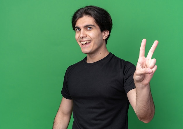 Foto grátis jovem bonito sorridente usando uma camiseta preta mostrando um gesto de paz isolado em uma parede verde com espaço de cópia