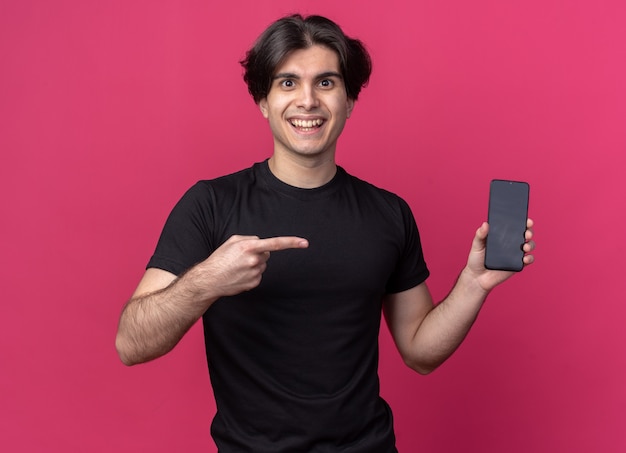 Jovem bonito sorridente usando uma camiseta preta segurando e apontando para o telefone isolado na parede rosa