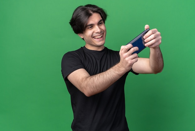 Jovem bonito sorridente usando uma camiseta preta tira uma selfie isolada em uma parede verde com espaço de cópia