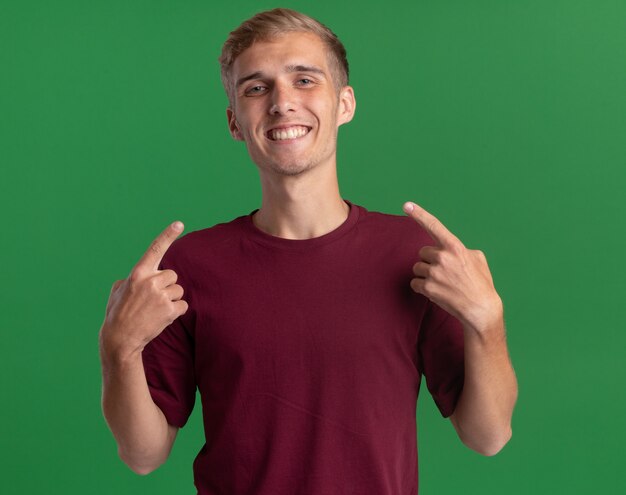 Jovem bonito sorridente, vestindo camisa vermelha, aponta para si mesmo, isolado na parede verde