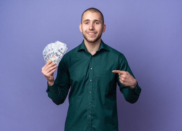 Jovem bonito sorridente, vestindo uma camisa verde segurando e apontando para o dinheiro