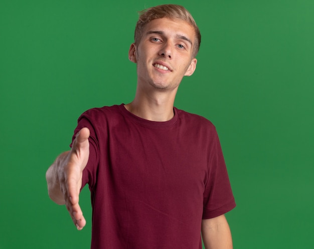 Foto grátis jovem bonito sorridente, vestindo uma camisa vermelha, estendendo a mão para a câmera, isolada na parede verde