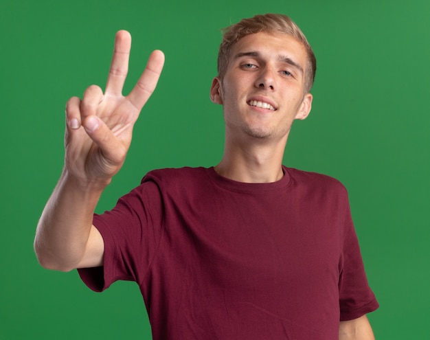 Jovem bonito sorridente, vestindo uma camisa vermelha, mostrando um gesto de paz isolado em uma parede verde