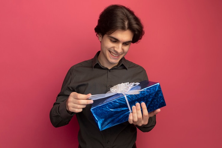 Foto grátis jovem bonito sorridente, vestindo uma camiseta preta, segurando e olhando para uma caixa de presente isolada na parede rosa