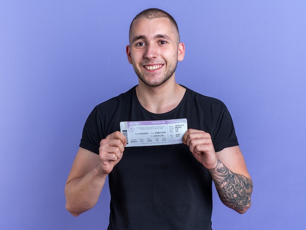 Jovem bonito sorridente, vestindo uma camiseta preta, segurando o ingresso isolado na parede azul