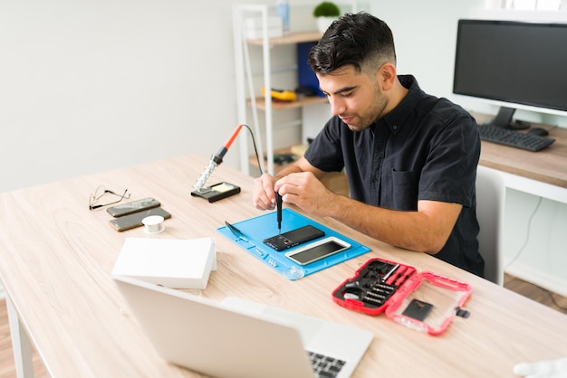 Jovem bonito sorrindo enquanto conserta um smartphone antigo. técnico masculino usando uma chave de fenda para consertar um celular quebrado na oficina