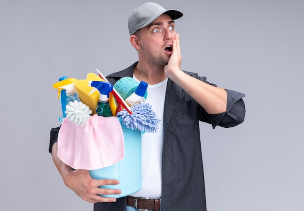 Jovem, bonito, surpreso, cara de limpeza vestindo camiseta e boné, segurando balde de ferramentas de limpeza, colocando a mão na bochecha isolada na parede branca