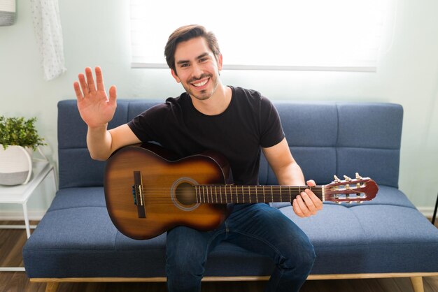 Jovem bonito tocando violão enquanto sorrindo e fazendo contato visual. Professor de música masculino pronto para seu aluno durante uma aula online