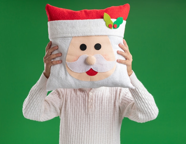 Foto grátis jovem bonito usando bandana de papai noel com óculos segurando uma almofada de papai noel na frente do rosto isolado sobre fundo verde