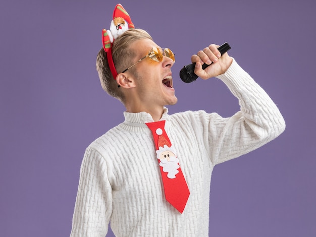 Jovem bonito usando bandana de papai noel e gravata com óculos segurando o microfone cantando com os olhos fechados, isolado em um fundo roxo com espaço de cópia