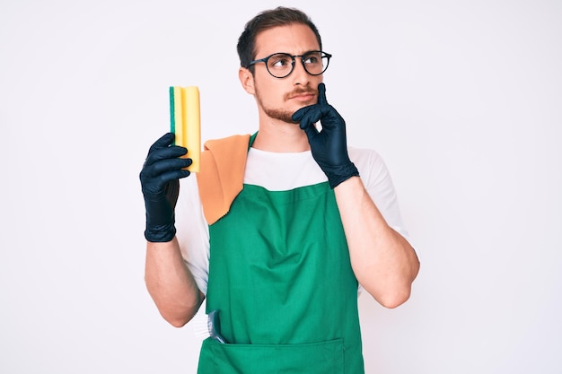 Foto grátis jovem bonito vestindo avental segurando esfregão rosto sério pensando em questão com a mão no queixo, pensativo sobre a ideia confusa