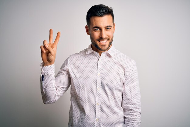 Jovem bonito vestindo camisa elegante em pé sobre fundo branco isolado sorrindo olhando para a câmera mostrando os dedos fazendo o sinal de vitória número dois