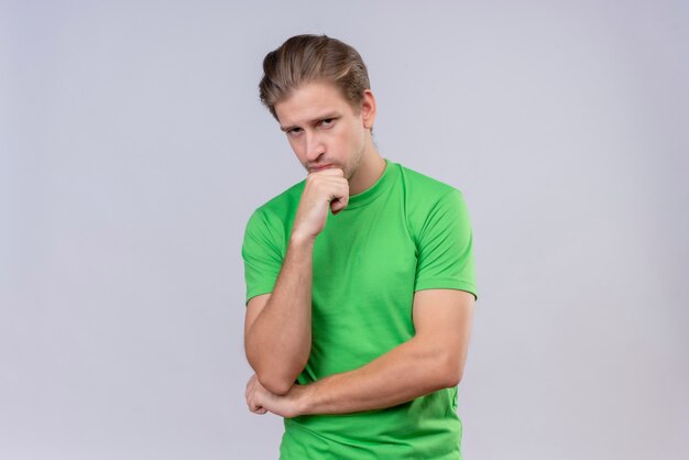 Jovem bonito vestindo camiseta verde com expressão pensativa no rosto e mão no queixo em pé sobre uma parede branca