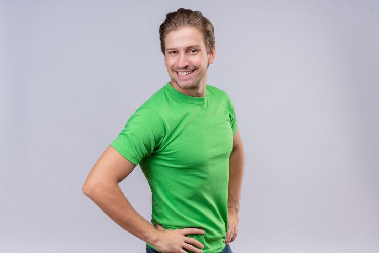 Foto grátis jovem bonito vestindo camiseta verde, de pé de lado com expressão confiante e satisfeito sobre a parede branca 2
