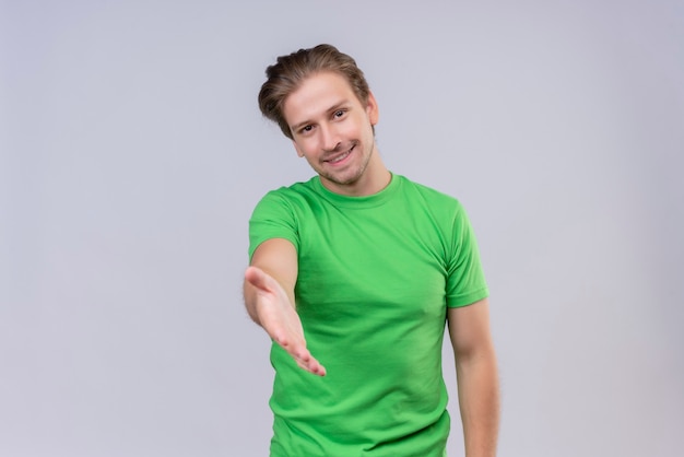 Jovem bonito vestindo camiseta verde e sorrindo amigável, fazendo gesto de saudação, oferecendo a mão em pé sobre uma parede branca