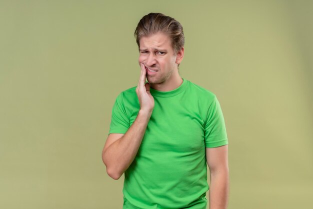 Jovem bonito vestindo camiseta verde parecendo doente tocando sua bochecha tendo dor de dente em pé sobre parede verde