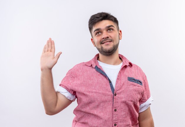 Jovem bonito vestindo uma camisa pólo rosa e dando minha palavra sinal com a mão esquerda levantada em pé sobre uma parede branca