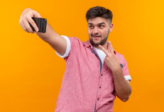 Jovem bonito vestindo uma camisa pólo rosa fazendo selfie mostrando o sinal da paz com os dedos em pé sobre a parede laranja