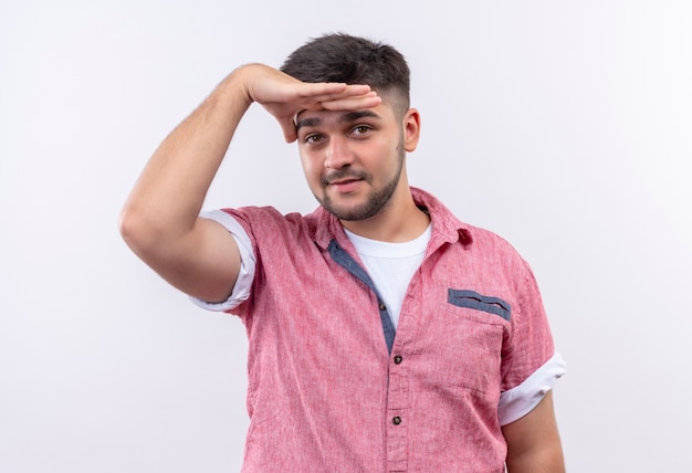 Foto grátis jovem bonito vestindo uma camisa pólo rosa olhando para a frente em pé sobre uma parede branca