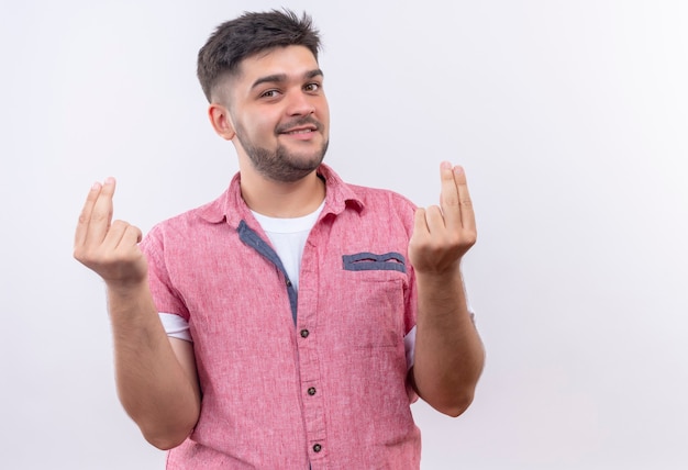 Jovem bonito vestindo uma camisa pólo rosa sorrindo mostrando o sinal do dinheiro com as mãos em pé sobre uma parede branca