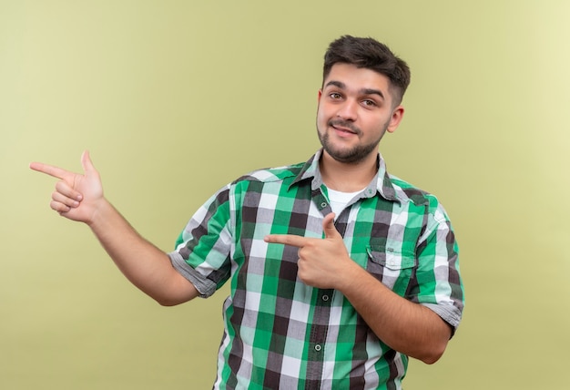 Jovem bonito vestindo uma camisa quadriculada, sorrindo, apontando para a direita com os indicadores em pé sobre a parede cáqui