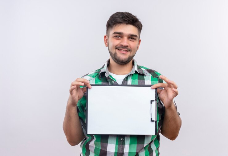 Foto grátis jovem bonito vestindo uma camisa quadriculada, sorrindo e segurando uma prancheta em pé sobre uma parede branca