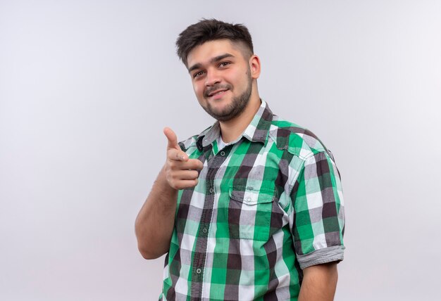 Jovem bonito vestindo uma camisa xadrez e sorrindo apontando em pé sobre uma parede branca