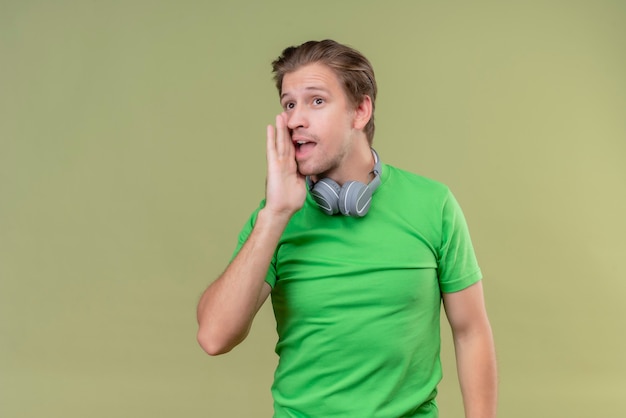 Jovem bonito vestindo uma camiseta verde com fones de ouvido contando um segredo com a mão perto da boca em pé sobre uma parede verde