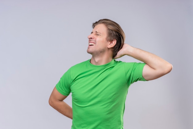 Jovem bonito vestindo uma camiseta verde parecendo indisposto e sentindo dores nas costas em pé sobre uma parede branca