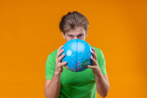 Jovem bonito vestindo uma camiseta verde segurando um globo escondido atrás dele com uma cara séria em pé sobre a parede laranja