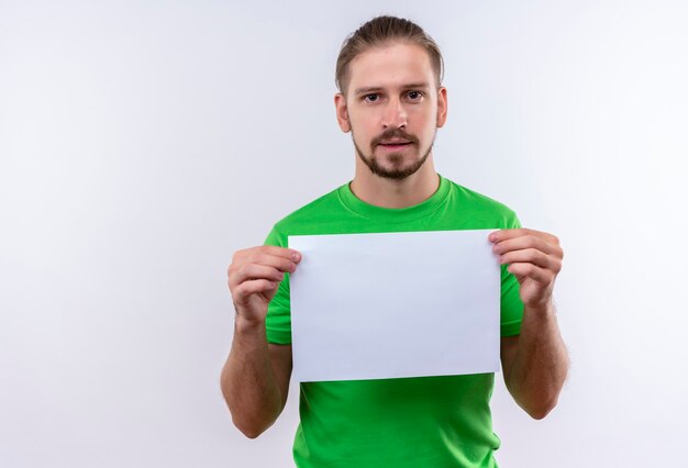 Jovem bonito vestindo uma camiseta verde segurando um papel em branco, olhando para a câmera, sorrindo confiante em pé sobre um fundo branco