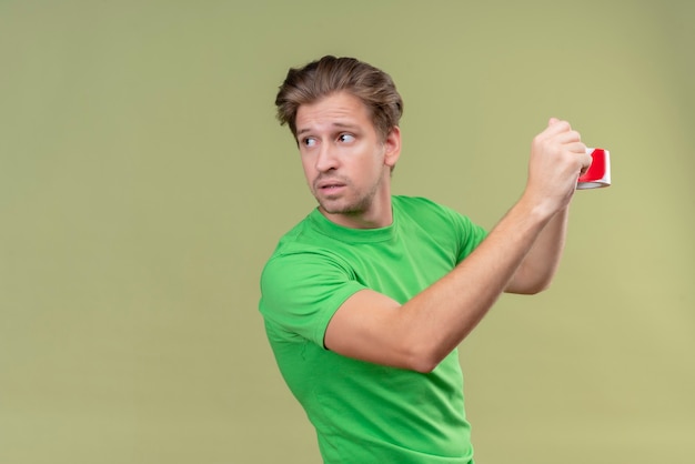 Jovem bonito vestindo uma camiseta verde usando fita adesiva parecendo confiante em pé sobre uma parede verde 5