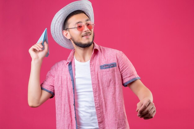 Jovem bonito viajante com chapéu de verão e óculos escuros, parecendo confiante jogando avião de papel em pé sobre fundo rosa